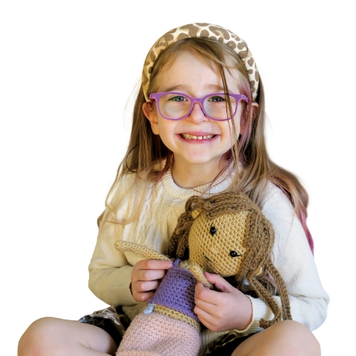 Portrait of Ivy, a 6-year-old smiling while holding a doll and wearing purple glasses against a light background